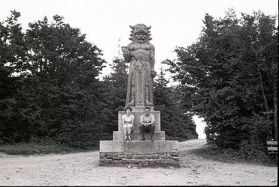 Monumental stone statue of a roaring lion-man hybrid atop a pedestal, flanked by two seated figures in 1930s attire. Dense fo...