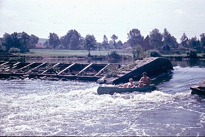 Vintage photo of two individuals in a small wooden boat navigating a rapid on a river, with a concrete dam spillway in the ba...