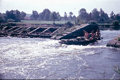Vintage photo of three individuals rafting on a rapid, using a makeshift raft of logs and tires. Concrete weir with stepped s...