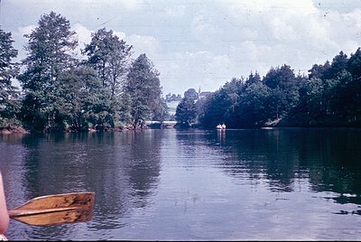 Vintage black-and-white boat on calm riverbank, surrounded by dense coniferous trees. Distant figures in water suggest rowing...