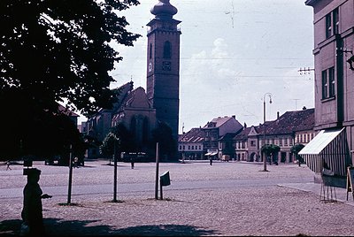 Vintage sepia-toned photo of a European town square, dominated by a tall clock tower with ornate details and a red-tiled chur...