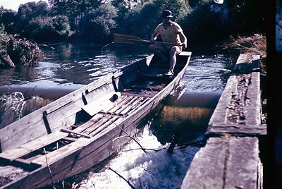 Vintage wooden boat with a seated fisherman rowing on a shallow river, surrounded by reeds and greenery. Mid-20th century rur...