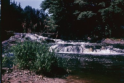 Vibrant 1970s-era waterfall cascading over layered rock formations into a calm pool, surrounded by dense foliage. Warm sepia-...