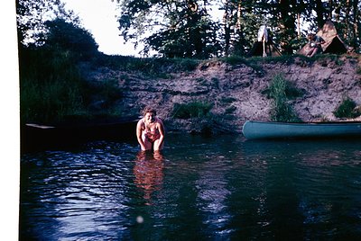 Vintage 1970s-style photograph of a woman standing in a shallow, natural river or lake. She wears a sleeveless top and shorts...