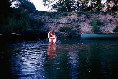 Vintage 1960s/70s outdoor scene: Couple embracing in shallow river, surrounded by lush greenery and rocky banks. Green canoe ...
