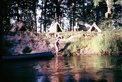 Vintage camping scene by a serene lake, featuring a woman in a swimsuit stepping from a canoe into shallow water. Wooden cano...