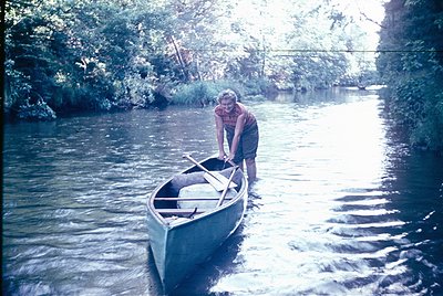 Vintage canoeing scene: man in 1970s-style plaid shirt and green pants prepares to launch a sleek, narrow canoe on a calm riv...