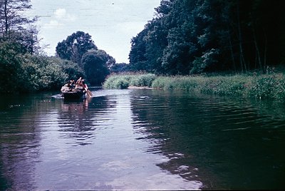 Vintage canoeing scene on a calm river flanked by dense greenery and mature trees. Four individuals in casual 1970s attire pa...