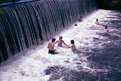 Vibrant 1960s/70s scene of children playing beneath a cascading waterfall. Concrete dam creates a natural pool; three kids in...