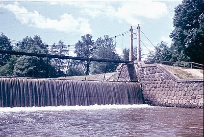 Vintage dam spillway with cascading water, flanked by stone retaining walls and utility poles. Mid-20th century engineering i...
