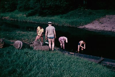 Four individuals in 1960s-style swimwear gather near a shallow, terraced waterway, likely for a communal dip or water activit...