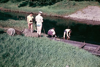 Four individuals in 1960s-style swimwear repair a wooden boat on a grassy riverside bank. The scene captures rural labor, lik...