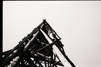 Black-and-white industrial shot of a wooden crane structure with a worker perched on the upper beam, likely mid-20th century....