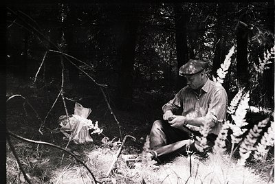 Black-and-white portrait of a man seated in dense foliage, holding a small object. His cap and rolled-sleeve attire suggest m...