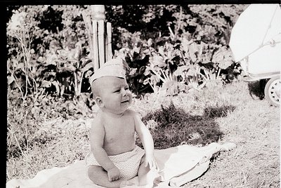 Mid-20th century black-and-white photo of a toddler in outdoor setting, wearing a sailor-style cap and diaper. Lush greenery ...