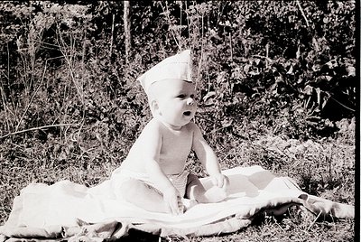 Mid-20th century black-and-white photo of an infant in a white diaper and paper crown, seated on a blanket outdoors among tal...