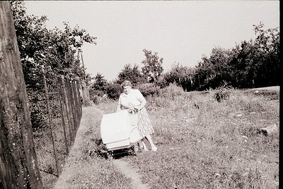 Black-and-white photo of a woman pushing a vintage lawnmower on a grassy path beside a rustic fence. Mid-20th century rural d...