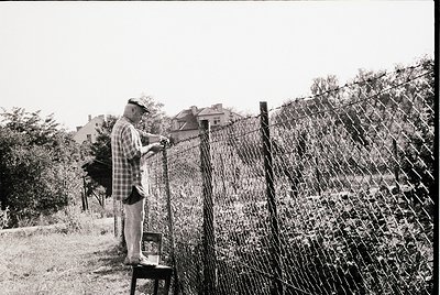 Mid-20th century man in plaid shirt and cap inspects barbed wire fence, likely during Cold War era. Urban/suburban residentia...