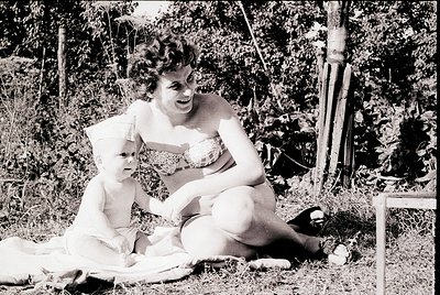 Black-and-white photo of a woman and child in outdoor setting, mid-20th century. Woman wears a two-piece swimsuit with decora...