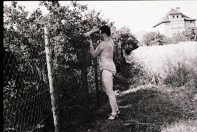 Black-and-white snapshot of a shirtless man in swim trunks adjusting sunglasses beside a dense hedge, mid-20th century. Rusti...