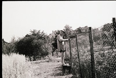 Child in plaid shirt leans over rusted chain-link fence, reaching toward a small, weathered wooden box on a stool. Overgrown ...