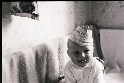 Vintage black-and-white photo of an infant wearing a newspaper hat and light-colored onesie, seated indoors. Wall displays ha...