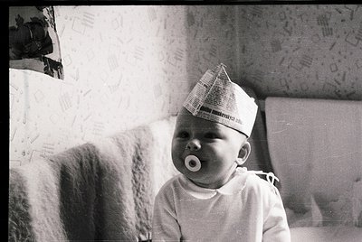 Vintage black-and-white photo of an infant wearing a cone-shaped paper hat (likely medical) and a pacifier, seated indoors on...