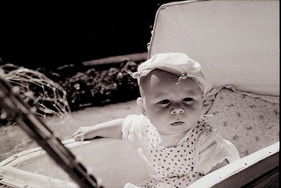 Vintage sepia-toned photo of a young child in a baby carriage, wearing a white bonnet and polka-dot dress. The carriage’s met...