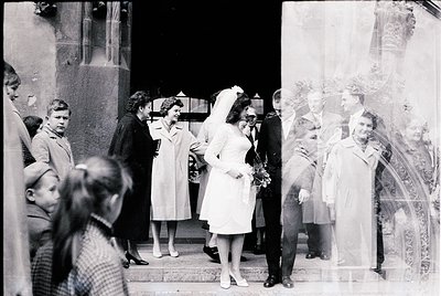 Vintage black-and-white wedding photo featuring a bride in a full-length white gown with a veil, exiting a grand building wit...