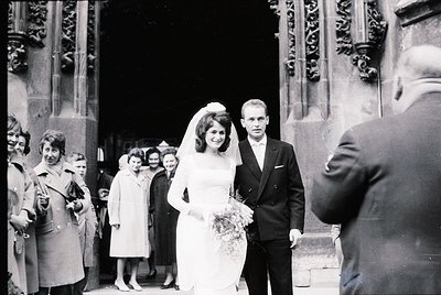1960s black-and-white wedding photo outside ornate church. Bride in full-length gown with veil, bouquet, and groom in dark su...