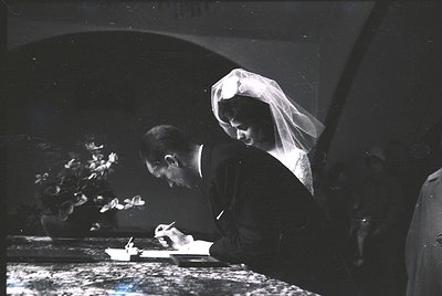 Black-and-white wedding ceremony shot featuring bride and groom signing a register. Bride wears veil and formal gown; groom i...