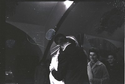 Vintage black-and-white photo of a couple dancing closely in a dimly lit room, likely a 1950s–1960s social event. The man’s a...