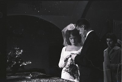 Mid-20th century black-and-white wedding portrait: bride in lace veil and formal gown, groom adjusting her veil, third person...