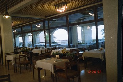 Mid-century seaside restaurant with arched windows overlooking ocean. Wooden tables draped in white linen, centerpiece flower...
