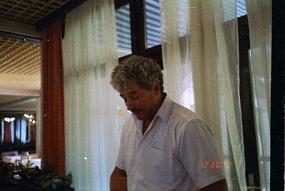 Mid-century man in a light-colored button-up shirt stands indoors near sheer curtains, framed by a wooden ceiling beam. Refle...
