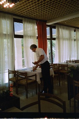 Mid-century dining hall with curved wooden benches, large windows draped with sheer curtains and red valances. A man in a whi...