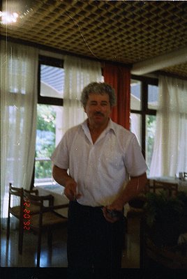 Mid-century indoor portrait of a man in a light button-down shirt and dark trousers, standing beside a wooden table with a di...