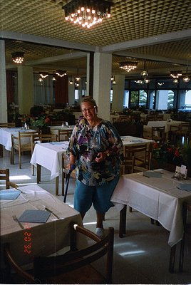 Elegant 1970s-style restaurant interior with wooden paneling, chandeliers, and neatly set tables. Woman in patterned blouse a...