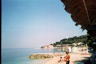 Vintage seaside scene with thatched roof shelter framing the shot. Rocky shoreline meets calm waters, lined with mid-20th-cen...