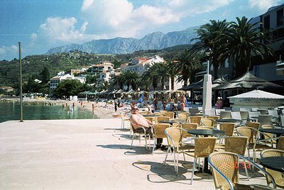 Seaside promenade with mid-century outdoor café featuring wicker chairs and tables under striped umbrellas. Mountainous backd...