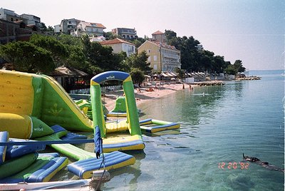 Waterfront scene featuring inflatable slides and loungers branded "wibit" along a sandy beach. Mid-20th century seaside resor...