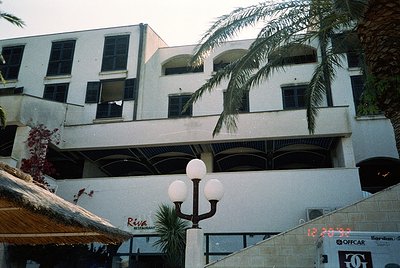 Mid-century seaside building with brutalist concrete balconies and arched windows, framed by palm trees. Signage includes "Ri...