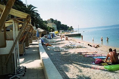 Sun-drenched seaside promenade with mid-20th-century beachgoers lounging on towels and sunbeds. Palm trees line the left, whi...