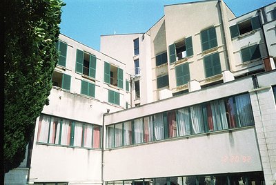 Mid-century Brutalist-style apartment block with concrete façade, large rectangular windows, and green shutters. Balconies fe...