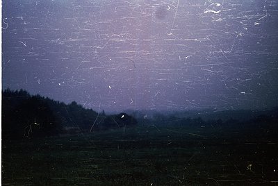 Vintage film grain overlay reveals a moody, rural landscape under storm clouds. Dark horizon silhouettes dense forest and dis...