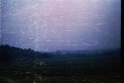 Vintage film grain overlay obscures a hazy, muted landscape with faint horizontal lines suggesting industrial or agricultural...