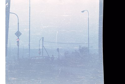 Urban street scene obscured by heavy fog, featuring blurred streetlights, traffic signals, and a pedestrian crossing sign. Th...