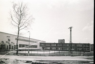 Industrial building with large glass-paneled facade, likely a factory or warehouse, framed by a chain-link fence. Bare tree a...