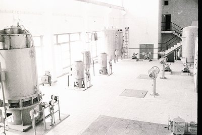 Industrial facility featuring mid-20th century processing equipment: large cylindrical tanks, conveyor belts, and mechanical ...