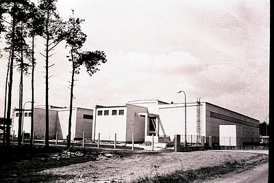 Mid-century modern industrial building with clean lines, flat roof, and large rectangular windows. Surrounded by tall pine tr...